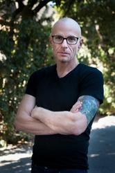 A man in a black t-shirt and glasses stands outside, with his arms crossed, looking directly at the camera.