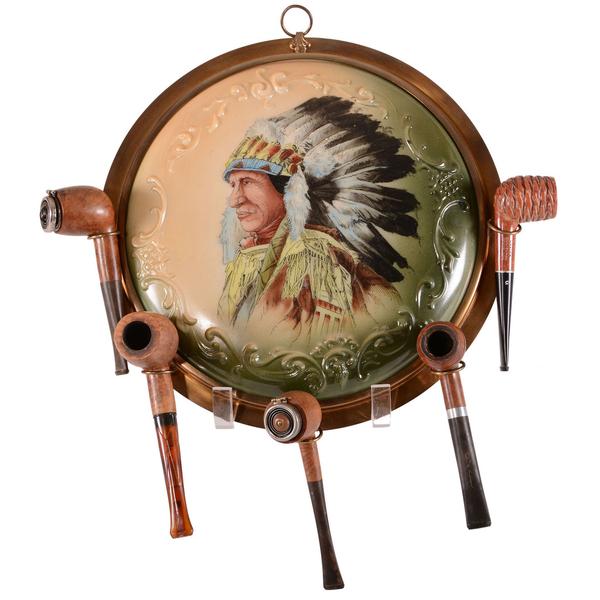 Unmarked Wave Crest pipe holder plaque in green and cream tones with a portrait of an Indian in full headdress, set in a brass frame holding five Wooden English pipes included in the lot ($13,000).