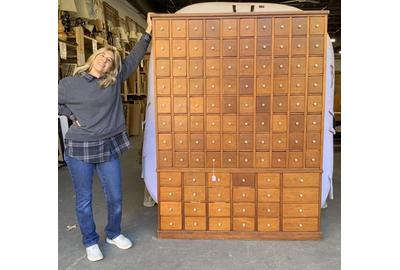 Pam Stone stands next to this rare and massive 104-drawer apothecary library card filing cabinet ($400-800) with hand dovetailing and porcelain knobs, which stands over 72 inches tall.