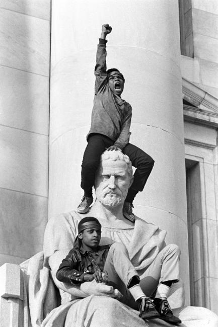  A boy giving a raised fist salute as he and a friend sat on a statue in front of the New Haven County Courthouse during the trial of Bobby Seale and Ericka Huggins.  