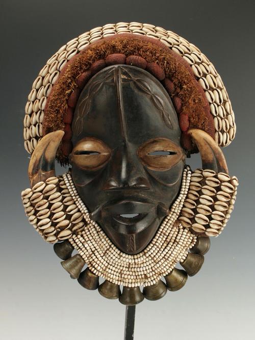 African Dan Guere mask, 23 inches by 10 inches, made from wood, wild boar horn, cowrie shells, beads, textiles, raffia and bronze bells, on a custom stand (est.  $300-$500).